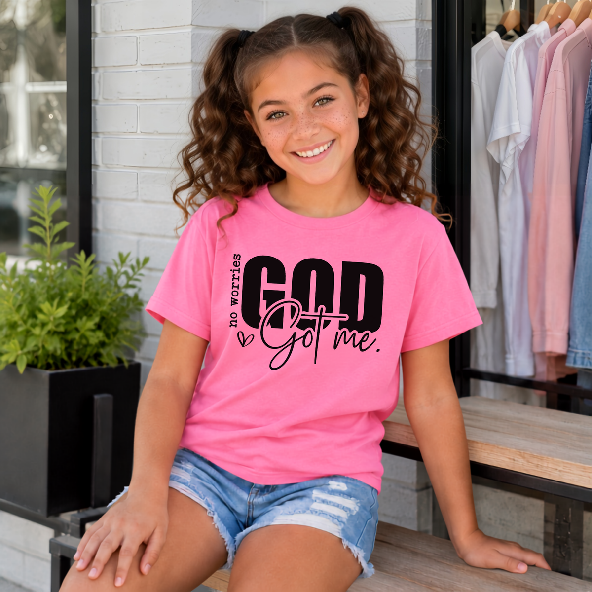Youth model wearing a bright pink No Worries God Got Me T-shirt with bold black lettering outside a storefront.