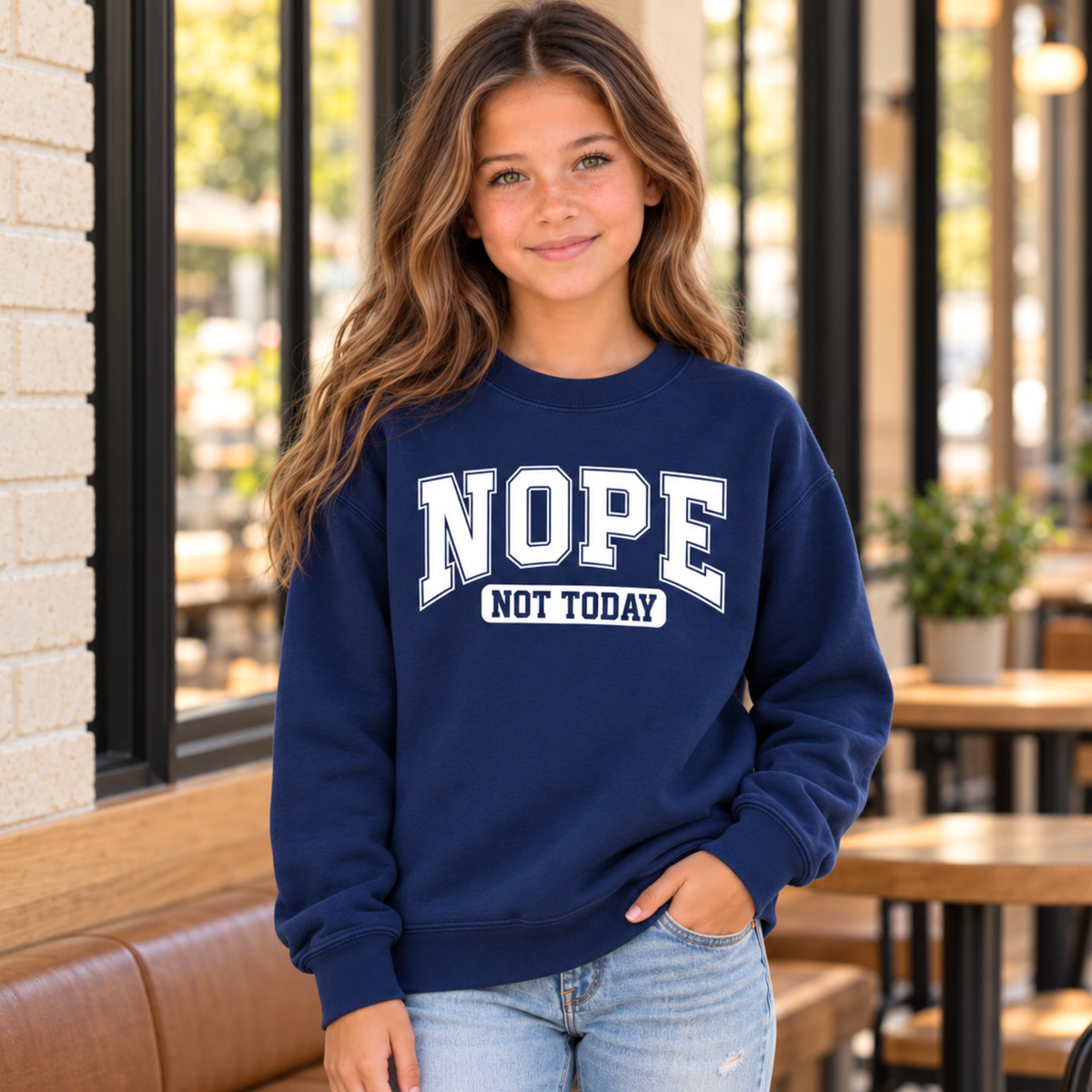 Navy youth crewneck sweatshirt with white NOPE NOT TODAY varsity lettering, modeled in a cafe.