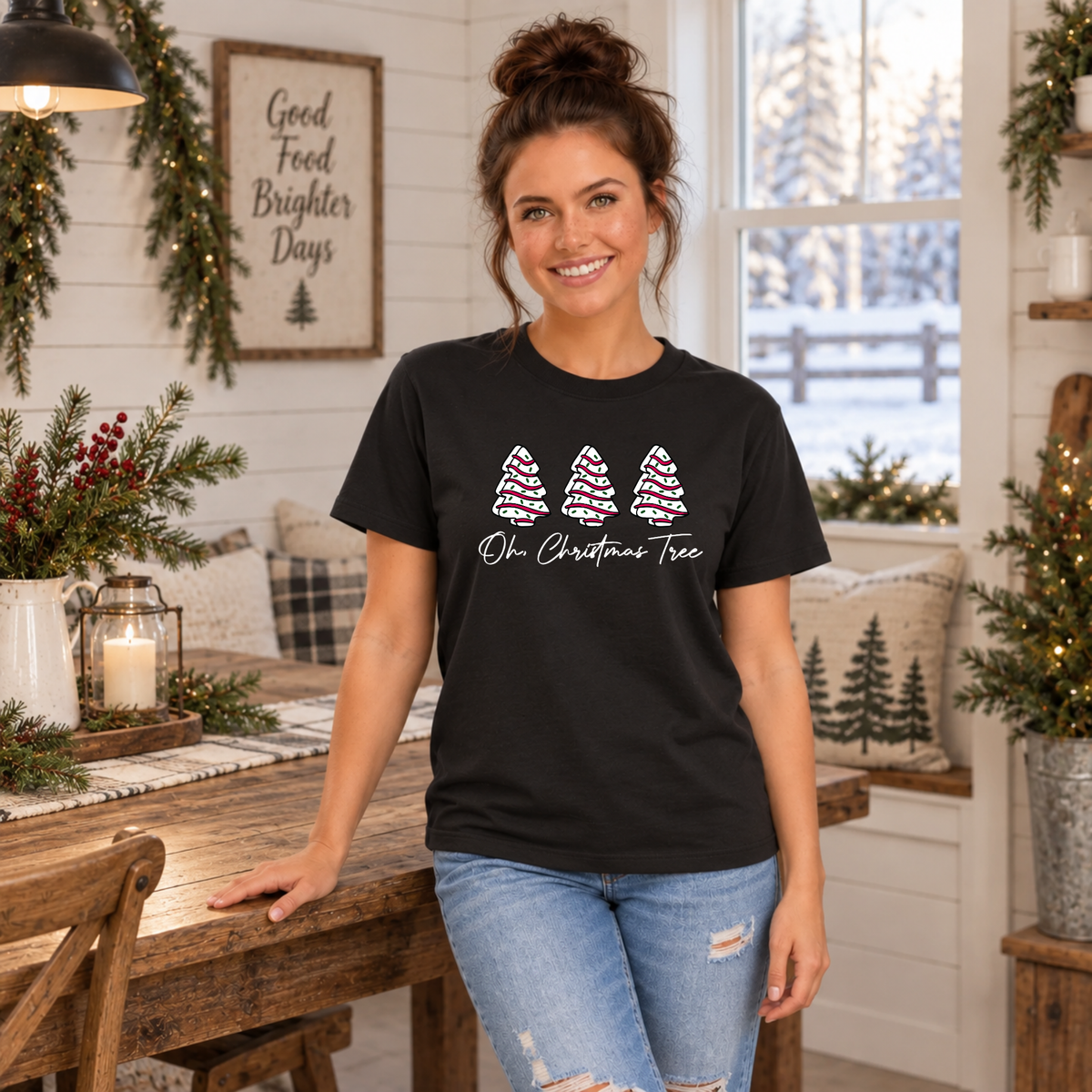 Woman in a black Oh Christmas Tree T-shirt leaning on a rustic dining table in a holiday farmhouse.