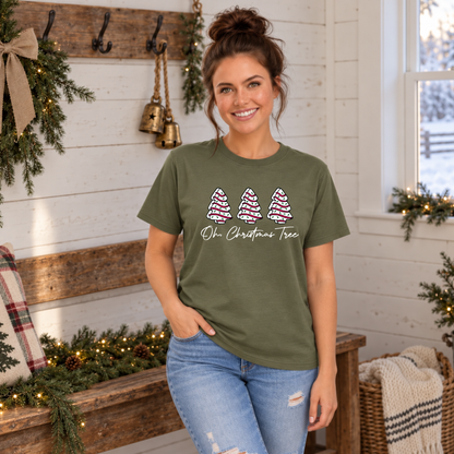 Woman in a military green Oh Christmas Tree T-shirt beside a wooden bench with garland and plaid pillow.