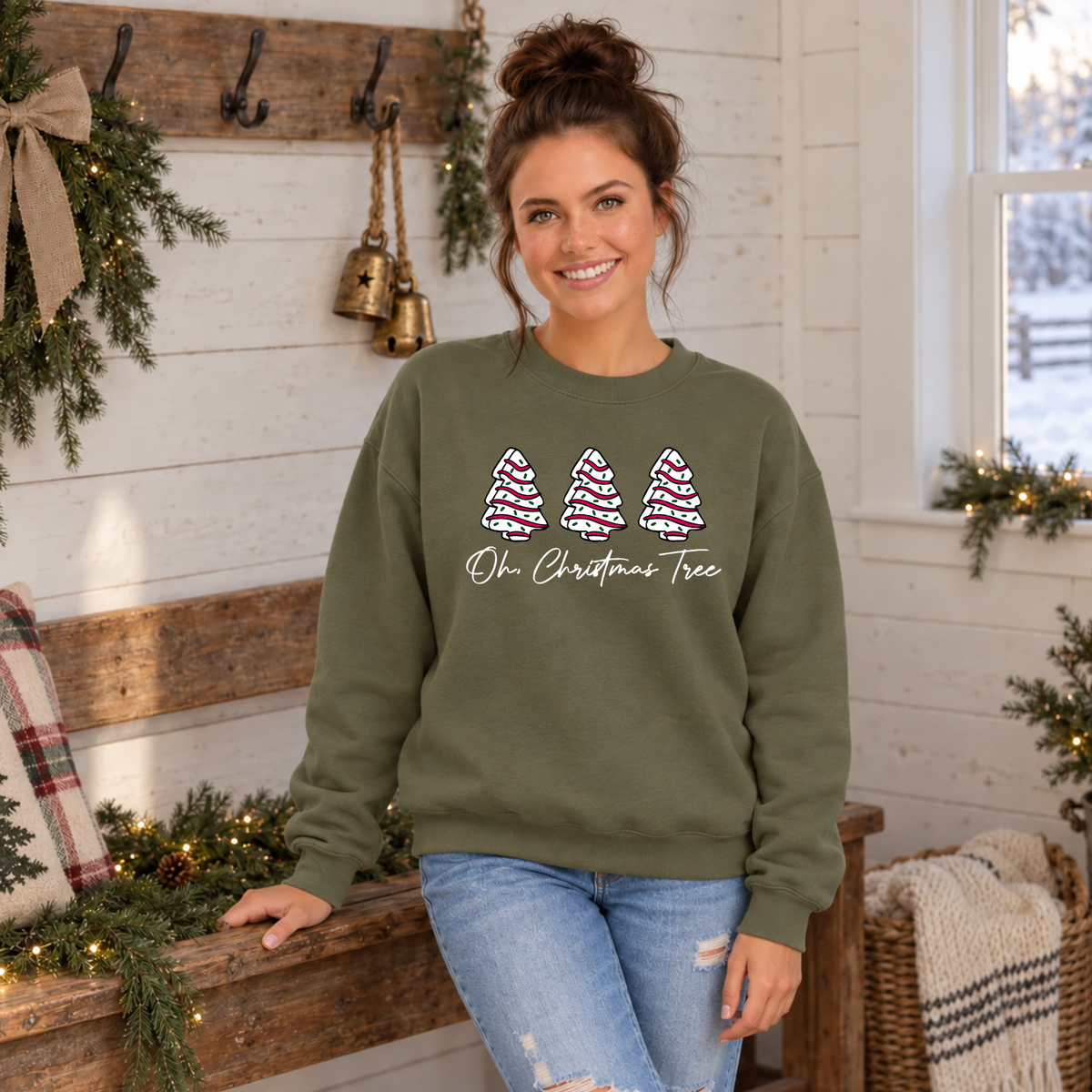 Woman wearing a military green Oh Christmas Tree sweatshirt beside a decorated bench and snowy window.