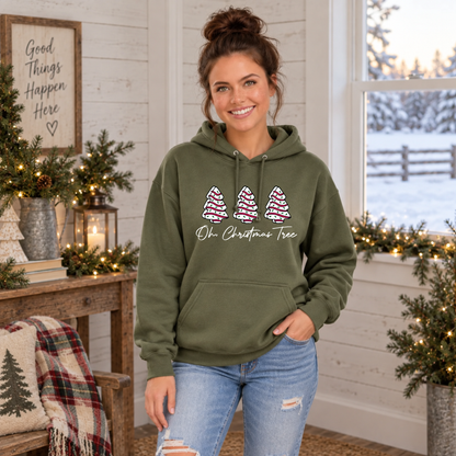 Woman wearing a military green Oh Christmas Tree hoodie in a cozy Christmas room with snow outside.