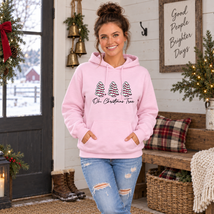 Woman wearing a light pink Oh Christmas Tree hoodie in a decorated entryway with a snowy farm outside.