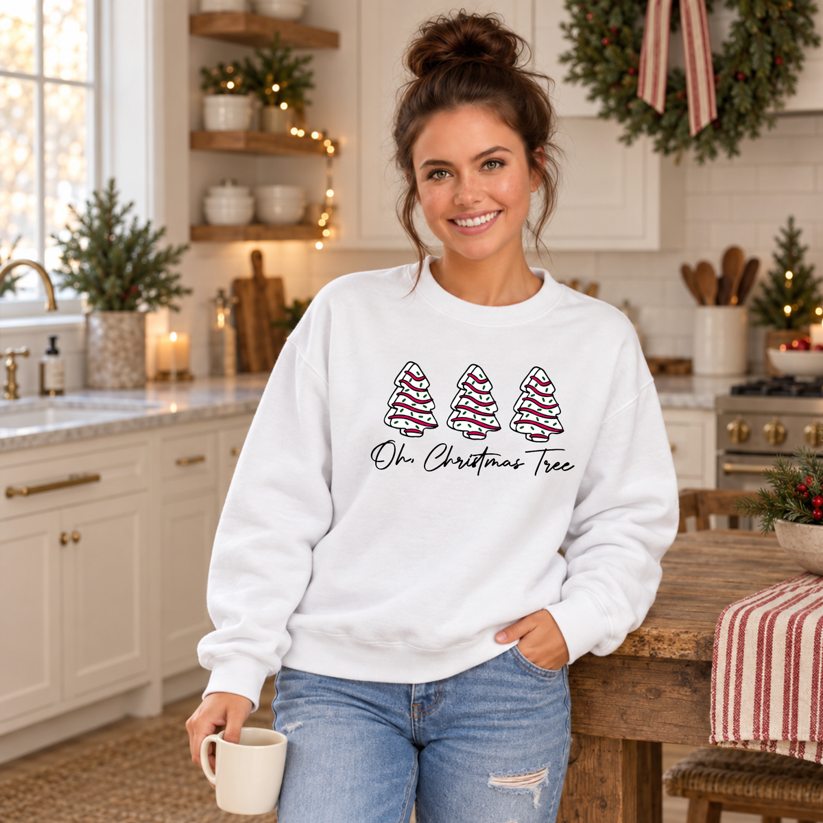 Woman in a white Oh Christmas Tree sweatshirt holding a mug in a Christmas kitchen.