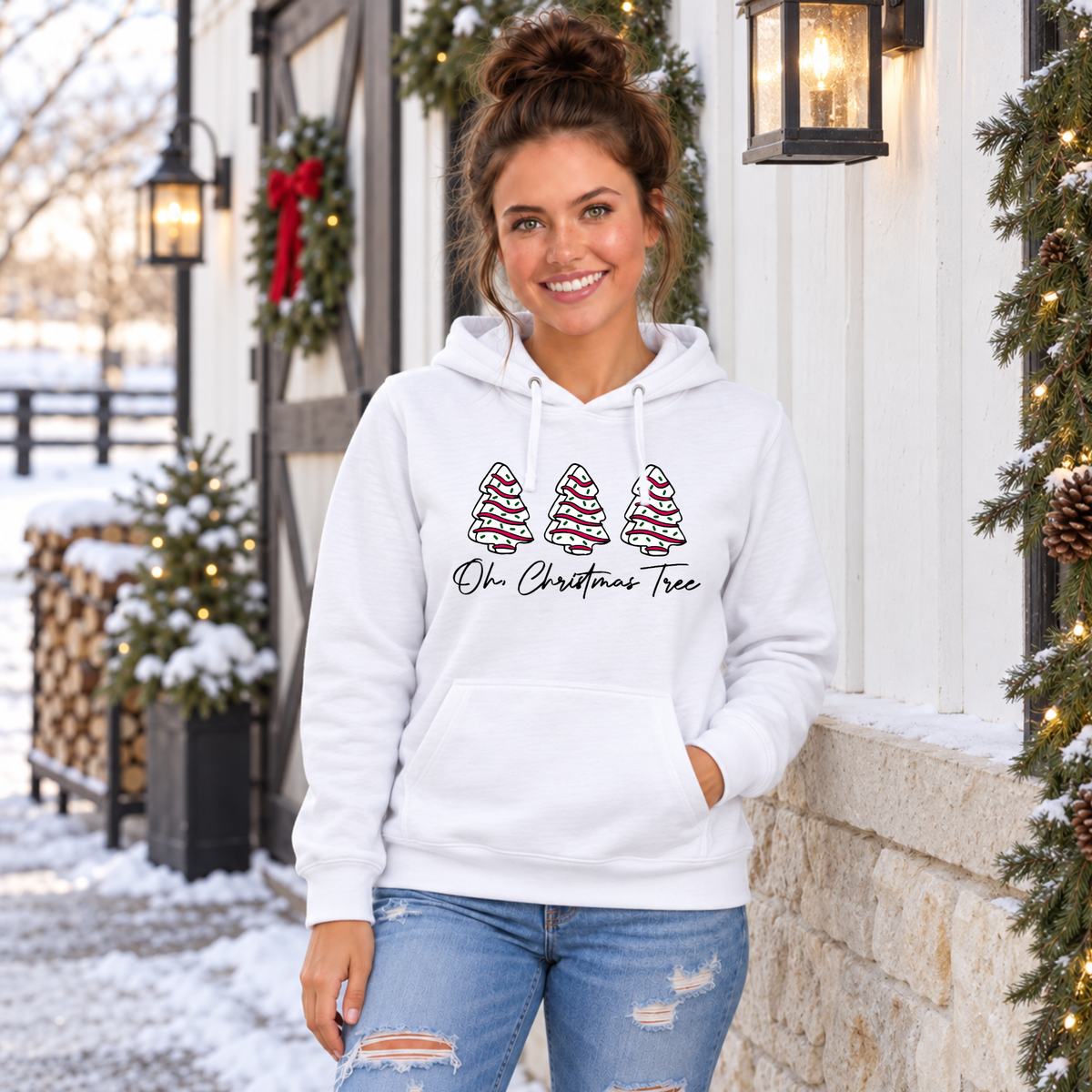 Woman in a white Oh Christmas Tree hoodie beside snow-covered firewood and a lit porch tree.