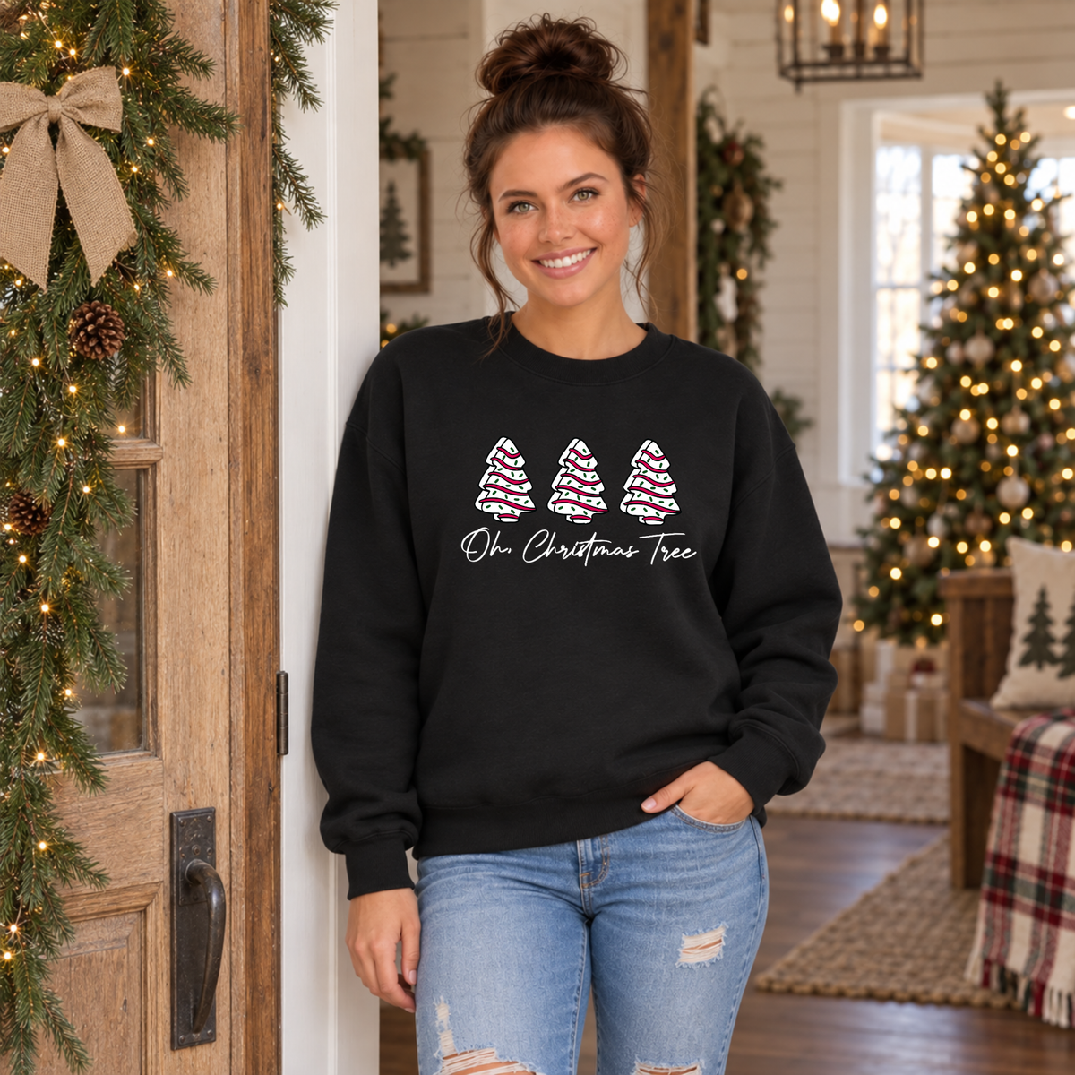 Woman wearing a black Oh Christmas Tree sweatshirt by a garland-trimmed door and lit Christmas tree.