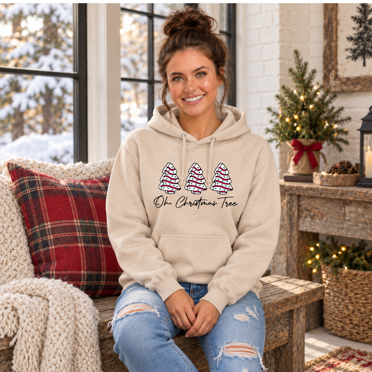 Woman in a sand Oh Christmas Tree hoodie seated by a plaid pillow and small lit Christmas tree.