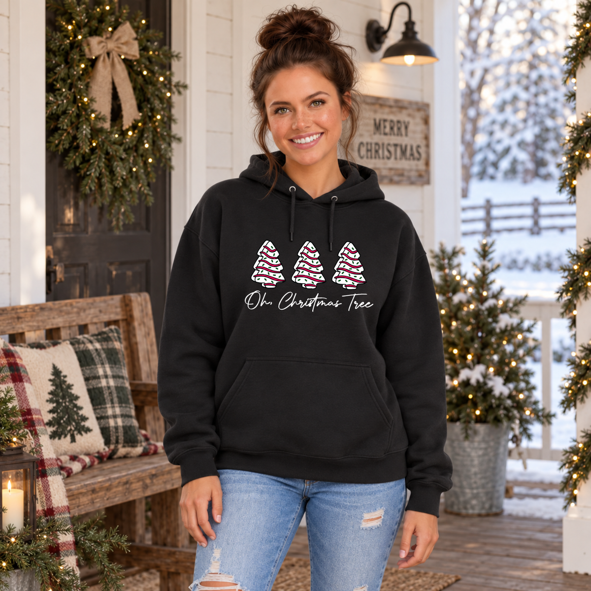 Woman in a black Oh Christmas Tree hoodie on a porch decorated with garland and a lit tree.