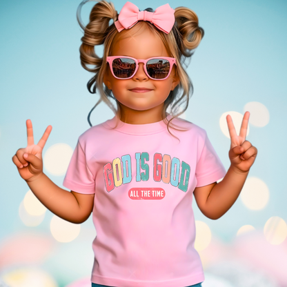 Smiling young girl showing peace sign and wearing a light pink T-shirt with the message 'God Is Good All the Time' in colorful text, symbolizing faith and positivity. Perfect for Easter and Christian-themed apparel.