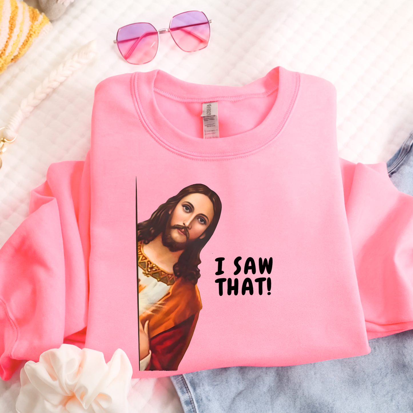 Pink sweatshirt with a graphic of a jesus saying  'I saw that!' text, placed on a textured surface.