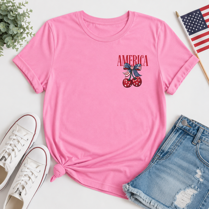Pink t-shirt with 'AMERICA' and cherry design, paired with white sneakers and denim shorts on a light surface.