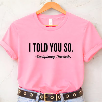Pink t-shirt with text 'I TOLD YOU SO. - Conspiracy Theorists' on a hanger.