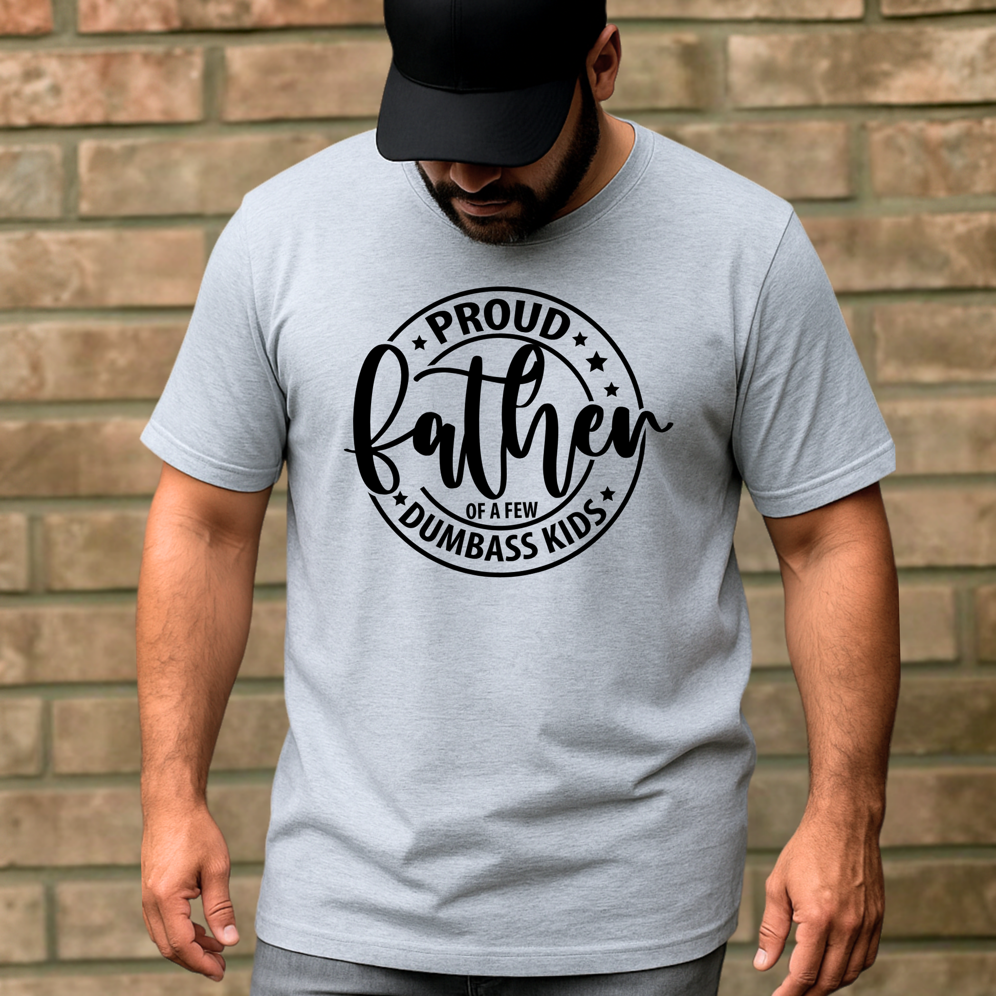Man wearing a gray t-shirt with 'Proud Father of a Few Dumbass Kids' text against a brick wall.