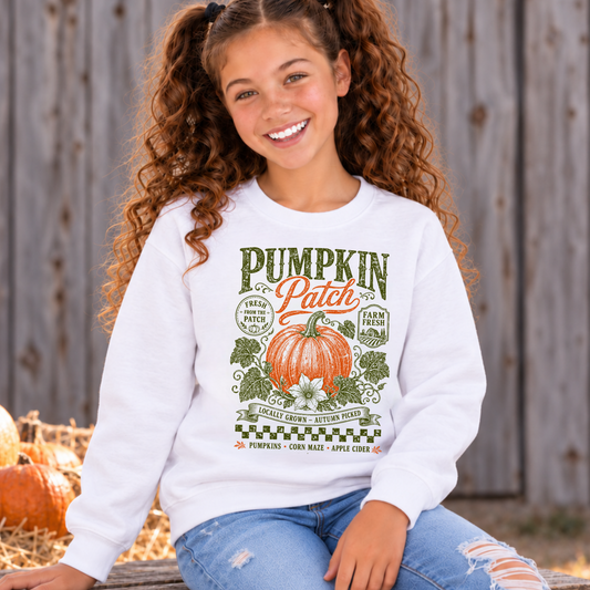 Girl wearing a white youth Pumpkin Patch sweatshirt while seated on a wooden bench at a rustic fall farm with pumpkins and a barn behind her