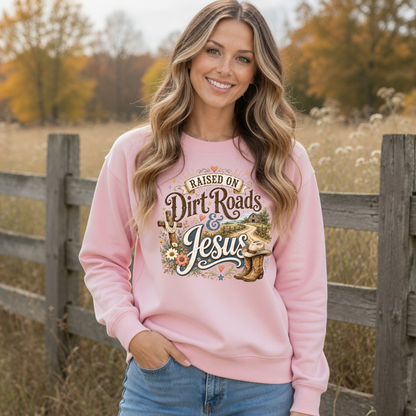 Woman modeling a light pink Raised on Dirt Roads & Jesus crewneck sweatshirt outdoors with a rustic fence and fall backdrop.