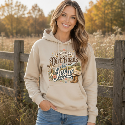 Woman wearing a sand Raised on Dirt Roads & Jesus hoodie outdoors in a golden autumn field.