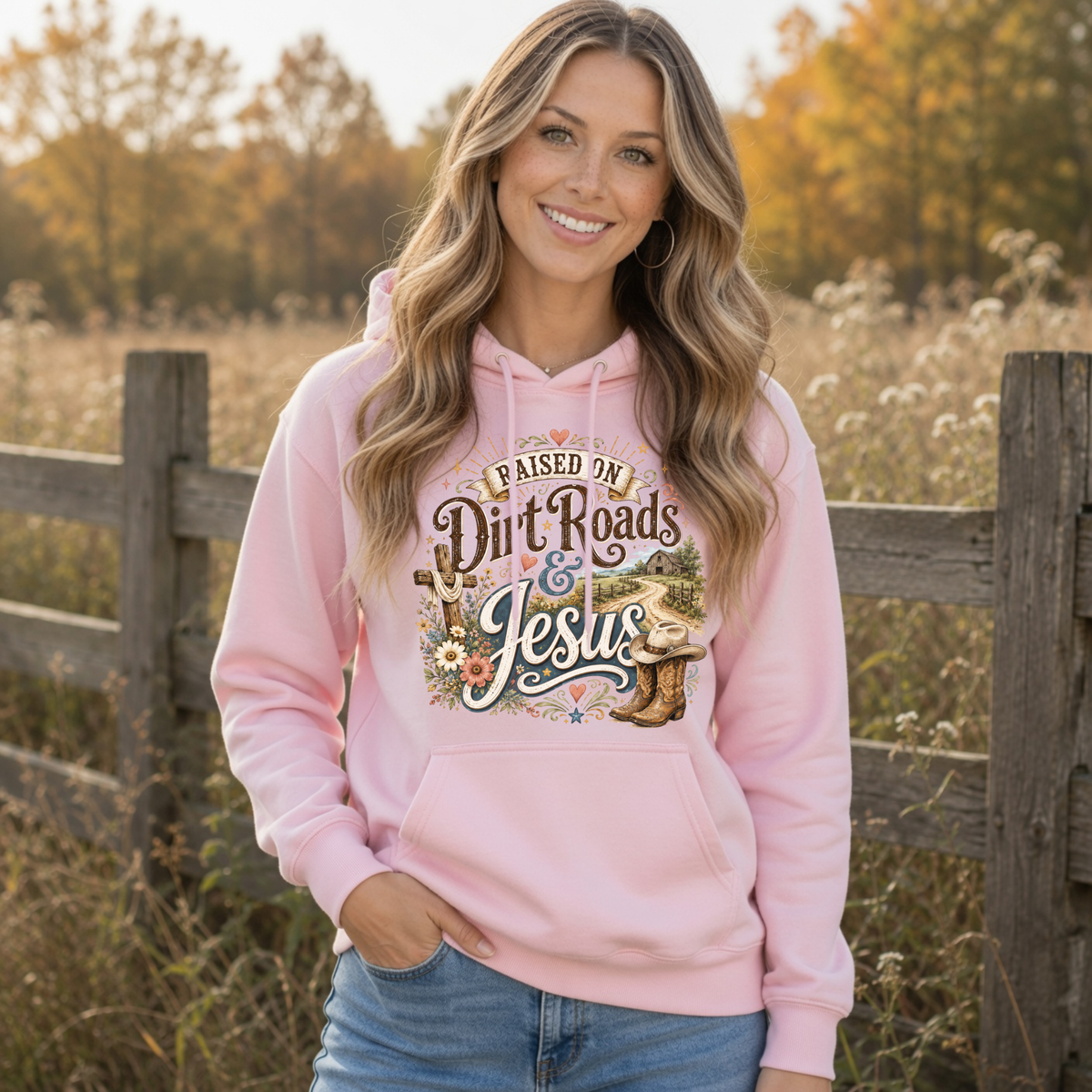 Woman modeling a light pink Raised on Dirt Roads & Jesus hoodie beside a wooden fence in a fall landscape.