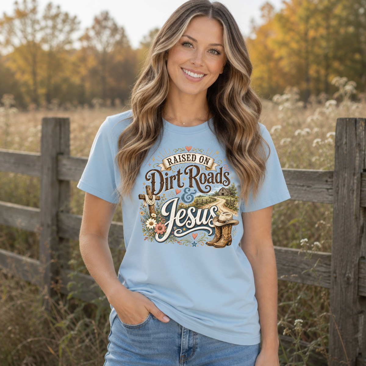 Woman wearing a light blue Raised on Dirt Roads & Jesus T-shirt outdoors with autumn foliage behind her.