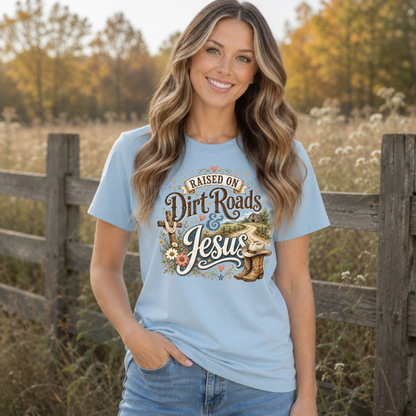 Woman wearing a light blue Raised on Dirt Roads & Jesus T-shirt outdoors with autumn foliage behind her.