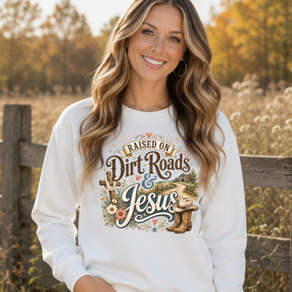 Woman wearing a white Raised on Dirt Roads & Jesus crewneck sweatshirt by a rustic fence in a fall field.