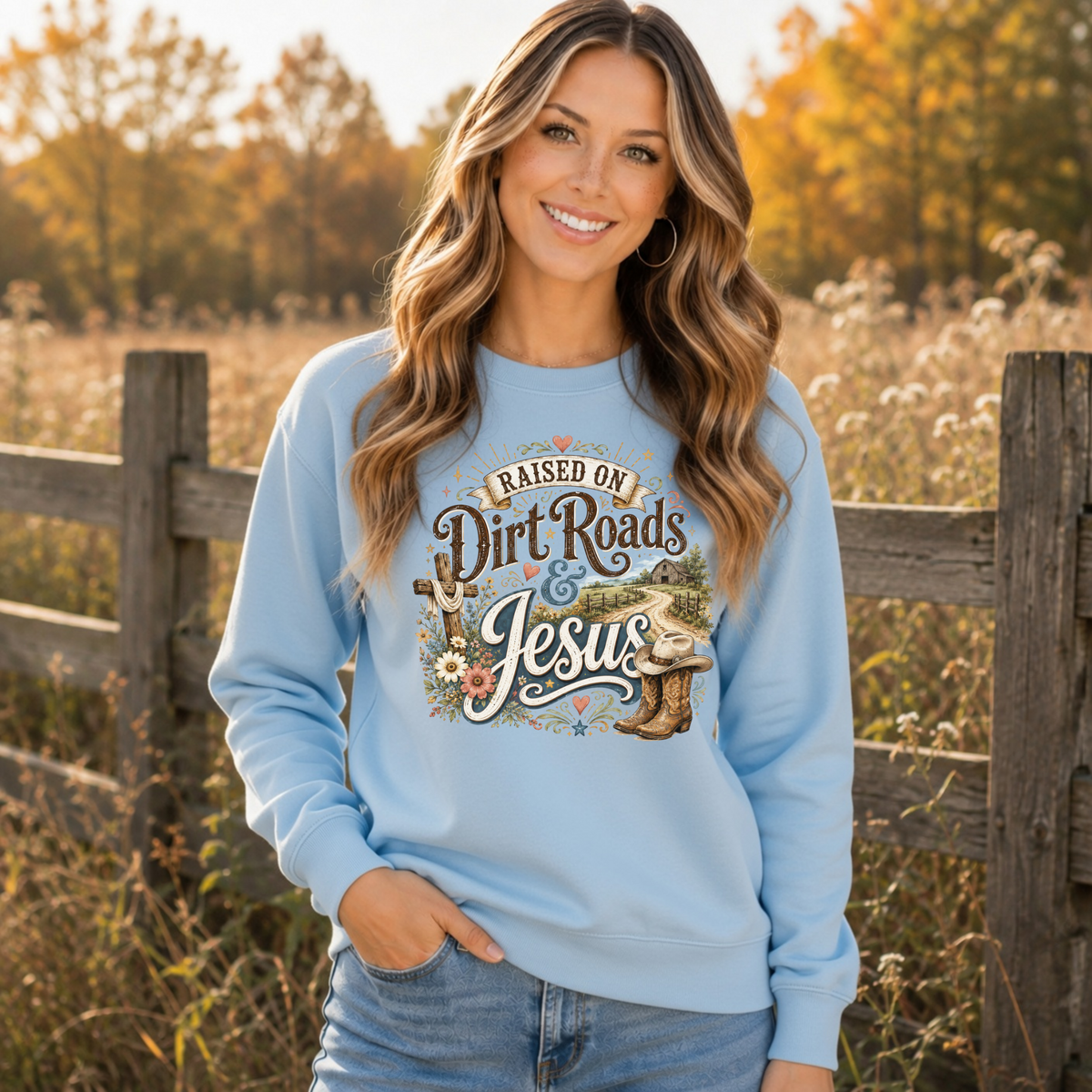 Woman wearing a light blue Raised on Dirt Roads & Jesus crewneck sweatshirt outdoors in an autumn country setting.