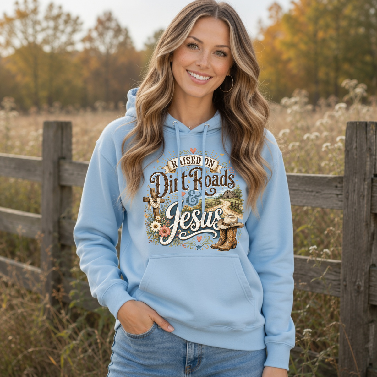 Woman modeling a light blue Raised on Dirt Roads & Jesus hoodie with a rustic fence and fall trees in the background.