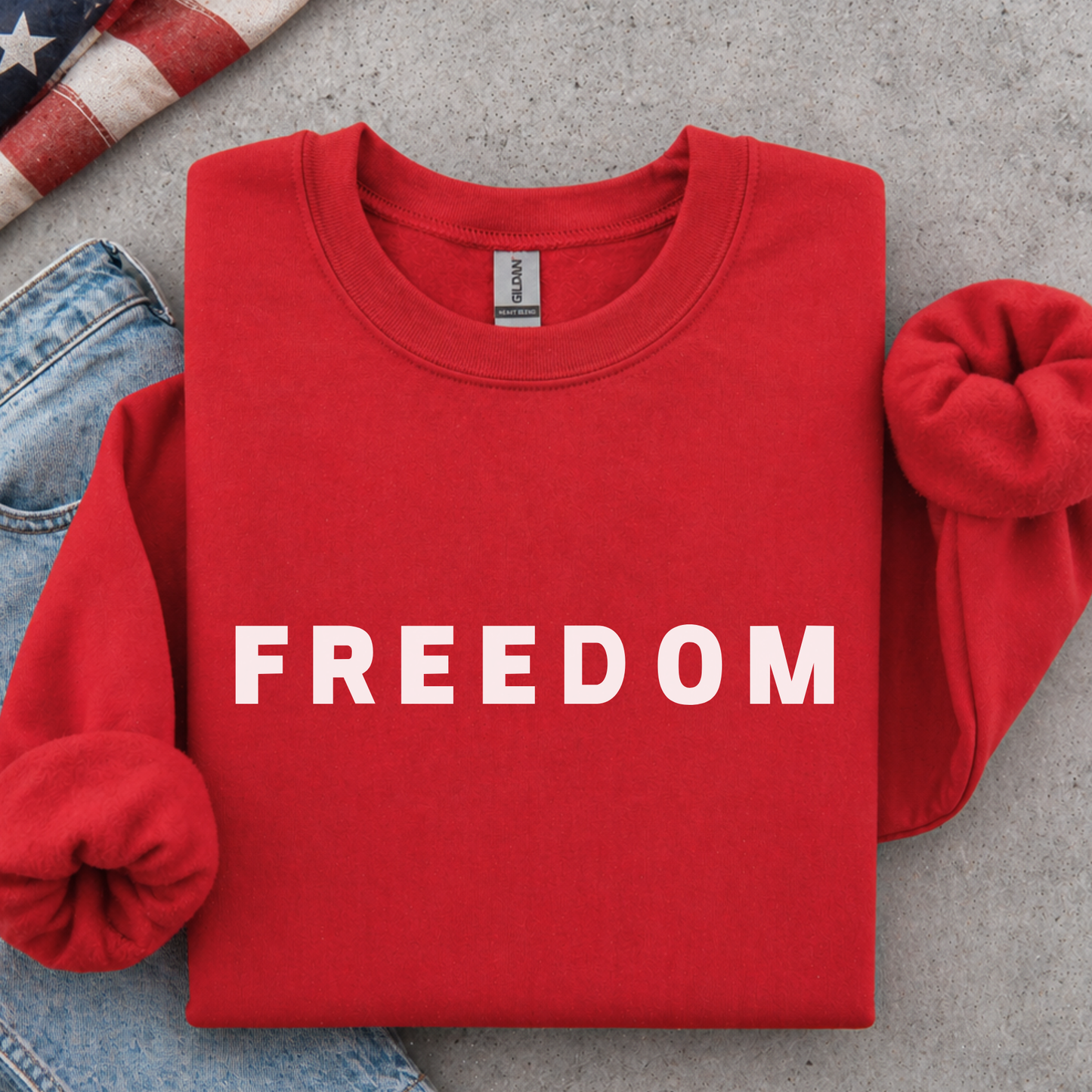 Red sweatshirt with 'FREEDOM' printed on it, placed on a concrete surface with jeans and an American flag in the background.