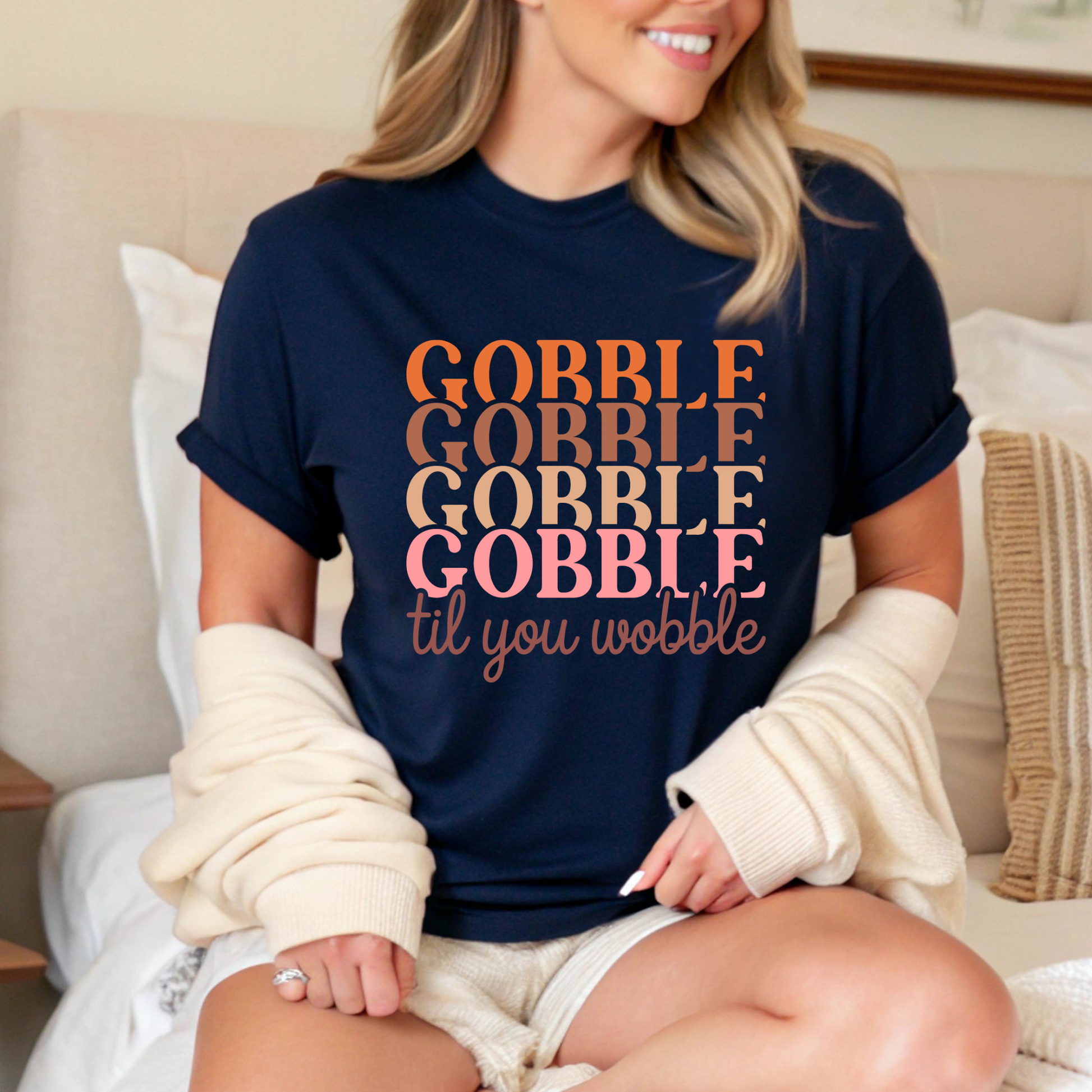 Relaxed woman in a navy blue shirt with “Gobble Til You Wobble” text, seated in a cozy bedroom setting wearing a warm cardigan