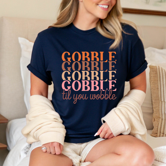 Relaxed woman in a navy blue shirt with “Gobble Til You Wobble” text, seated in a cozy bedroom setting wearing a warm cardigan