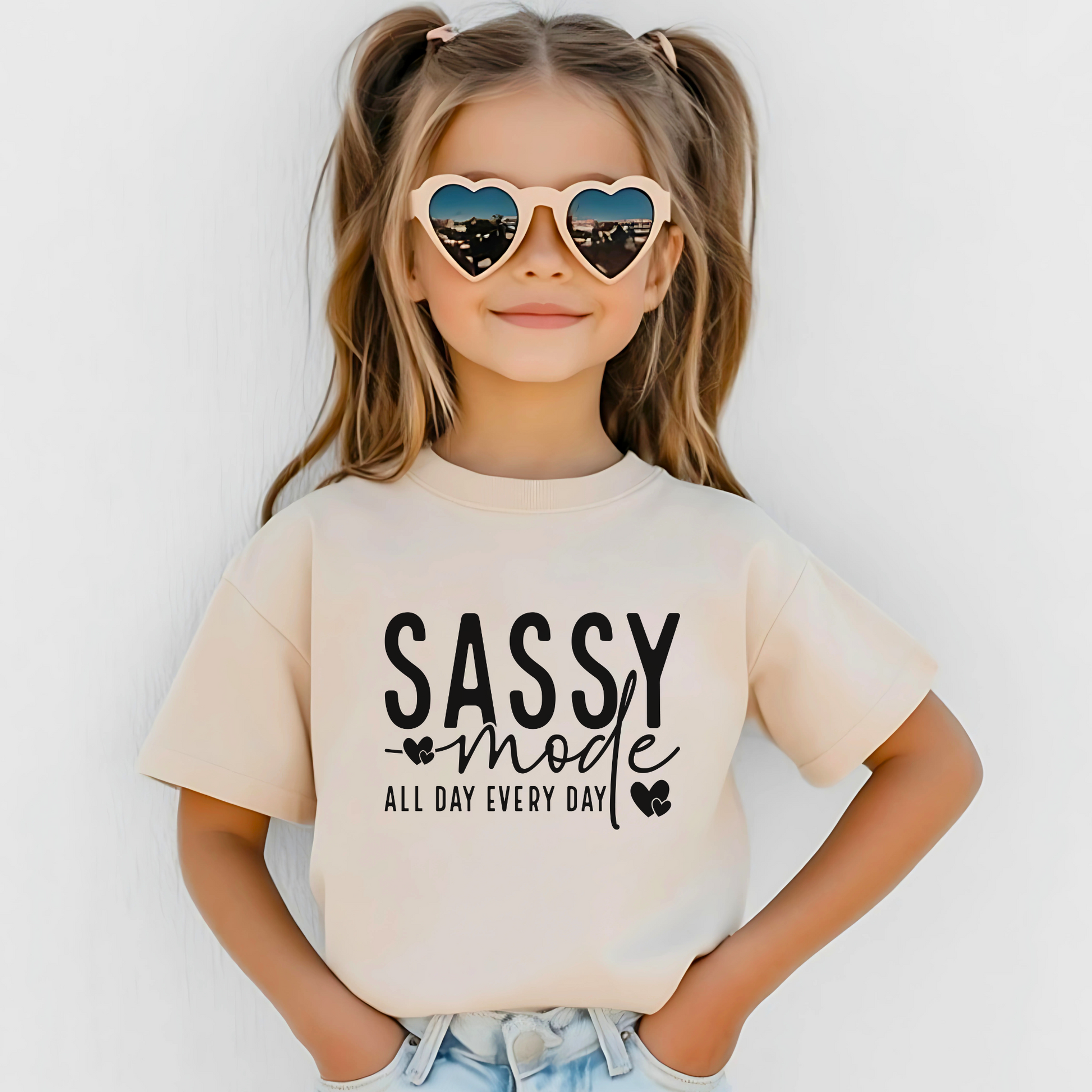 Young girl wearing a Sand "Sassy Mode – All Day Every Day" graphic t-shirt, smiling confidently outdoors