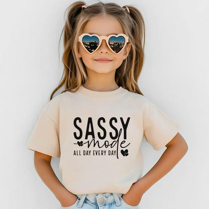 Young girl wearing a Sand "Sassy Mode – All Day Every Day" graphic t-shirt, smiling confidently outdoors