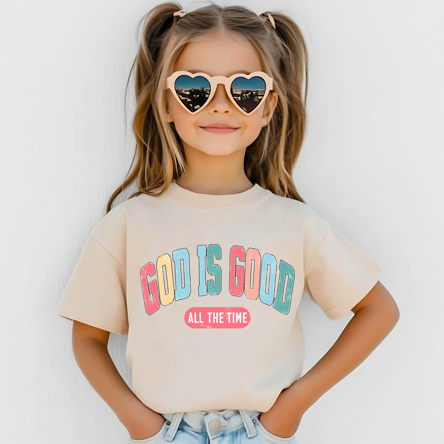 Smiling girl wearing a tan T-shirt with the message 'God Is Good All the Time' in colorful text, symbolizing faith and positivity. Perfect for Easter and Christian-themed apparel.