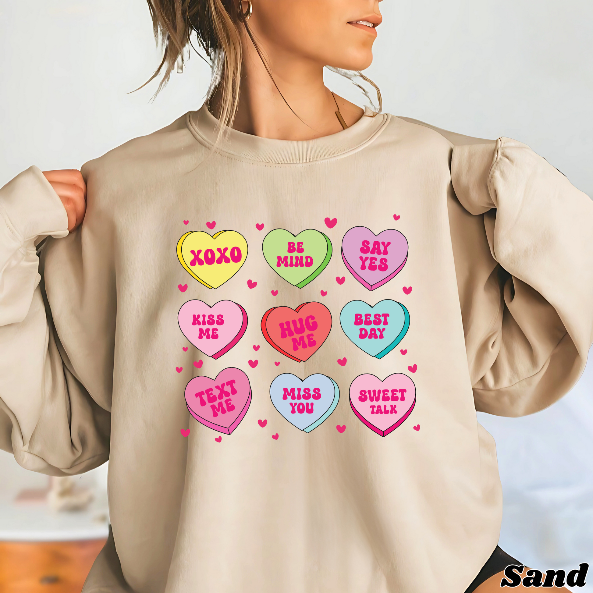 Woman wearing a sand Valentine’s sweatshirt featuring colorful candy conversation hearts with messages like “XOXO,” “Be Mine,” “Say Yes,” “Kiss Me,” “Hug Me,” and more.