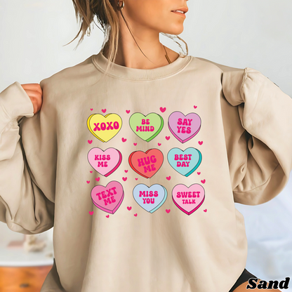 Woman wearing a sand Valentine’s sweatshirt featuring colorful candy conversation hearts with messages like “XOXO,” “Be Mine,” “Say Yes,” “Kiss Me,” “Hug Me,” and more.