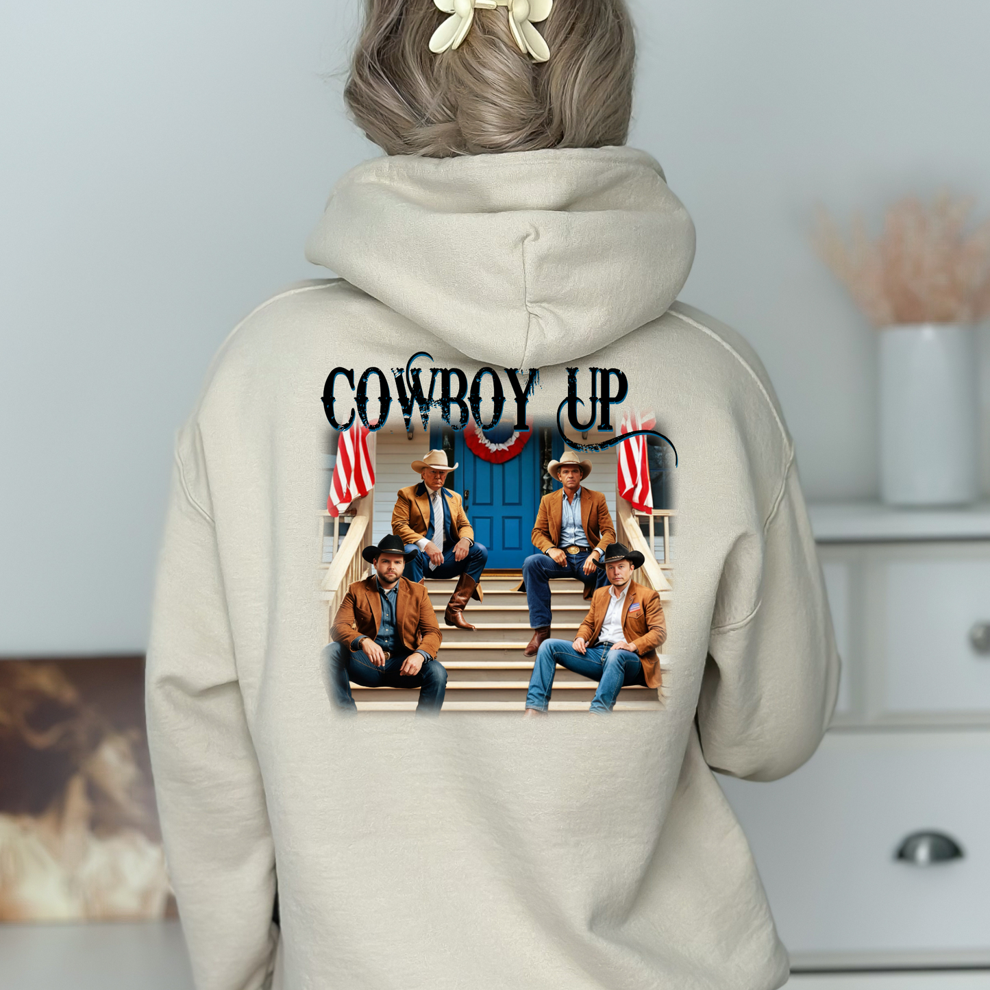 A white hoodie with a graphic print of cowboys seated on a porch with American flags and the text 'Cowboy Up' on the back and a front-left chest portrait of a cowboy with 'Cowboy Up' script.