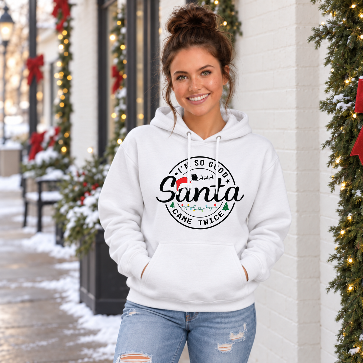 Woman wearing a white hoodie with I'm So Good Santa Came Twice Christmas design outside a snowy holiday storefront