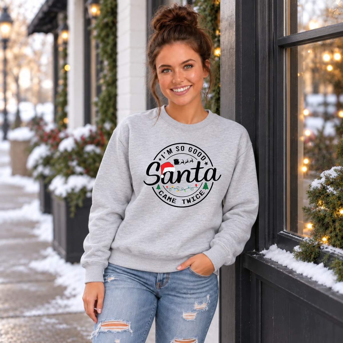 Woman wearing a sport gray sweatshirt with I'm So Good Santa Came Twice Christmas design outside a snowy holiday storefront