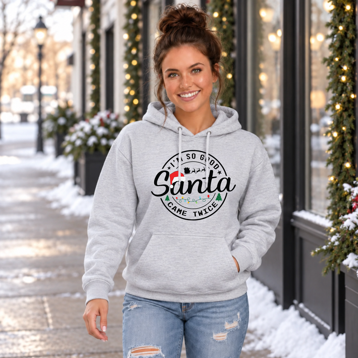 Woman wearing a sport gray hoodie with I'm So Good Santa Came Twice Christmas design outside a snowy decorated storefront