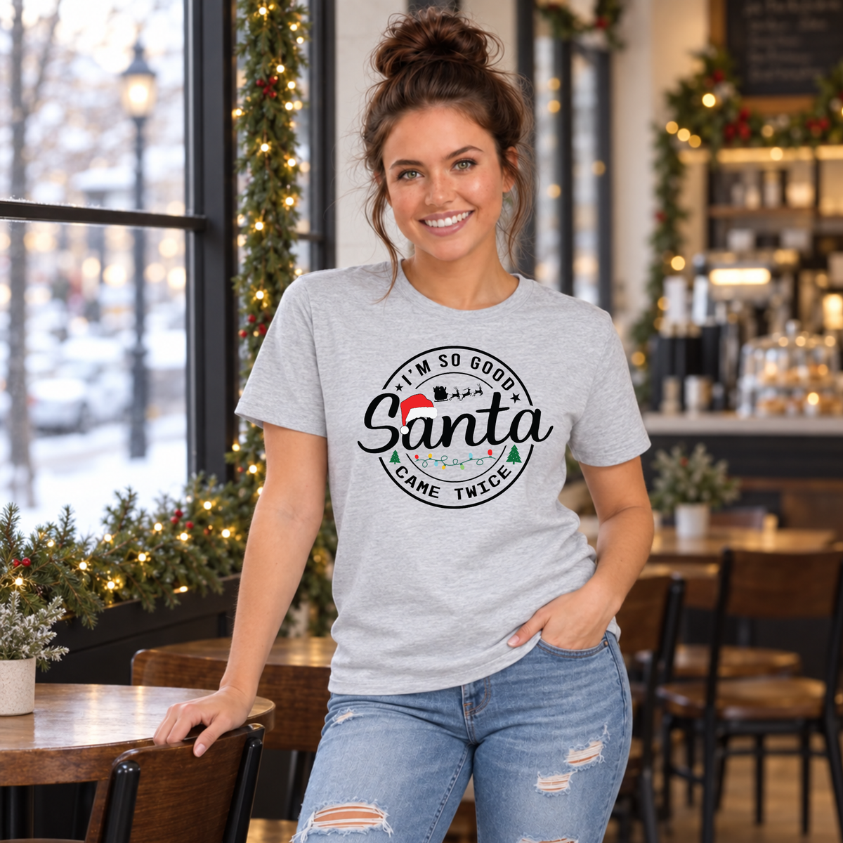 Woman wearing a sport gray T-shirt with I'm So Good Santa Came Twice Christmas design in a decorated café