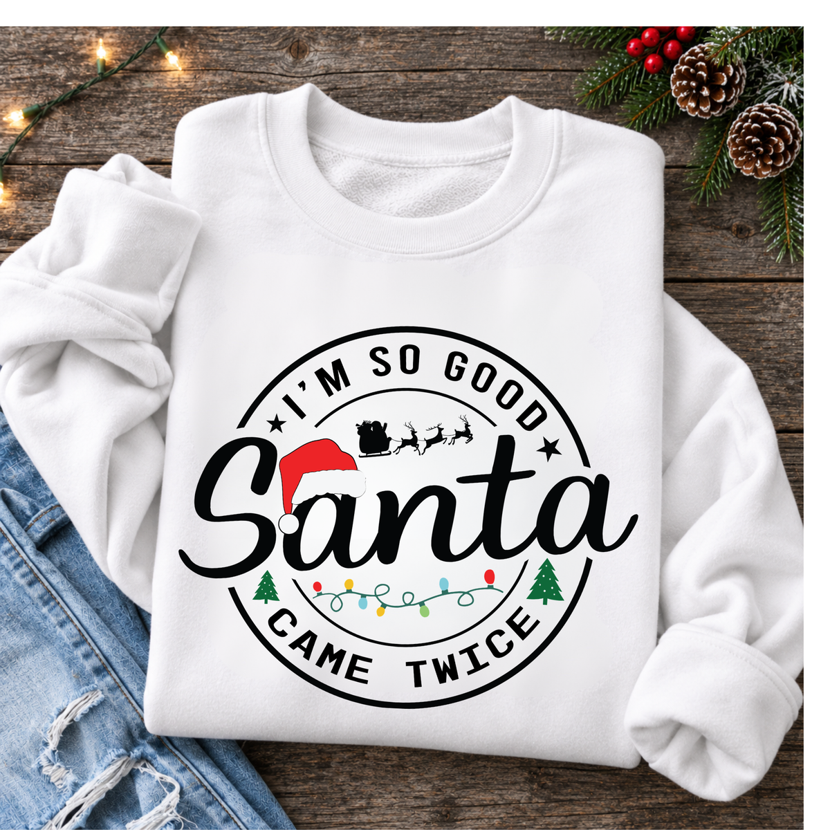 White sweatshirt flat lay with I'm So Good Santa Came Twice Christmas design, jeans, evergreens, pinecones and holiday lights