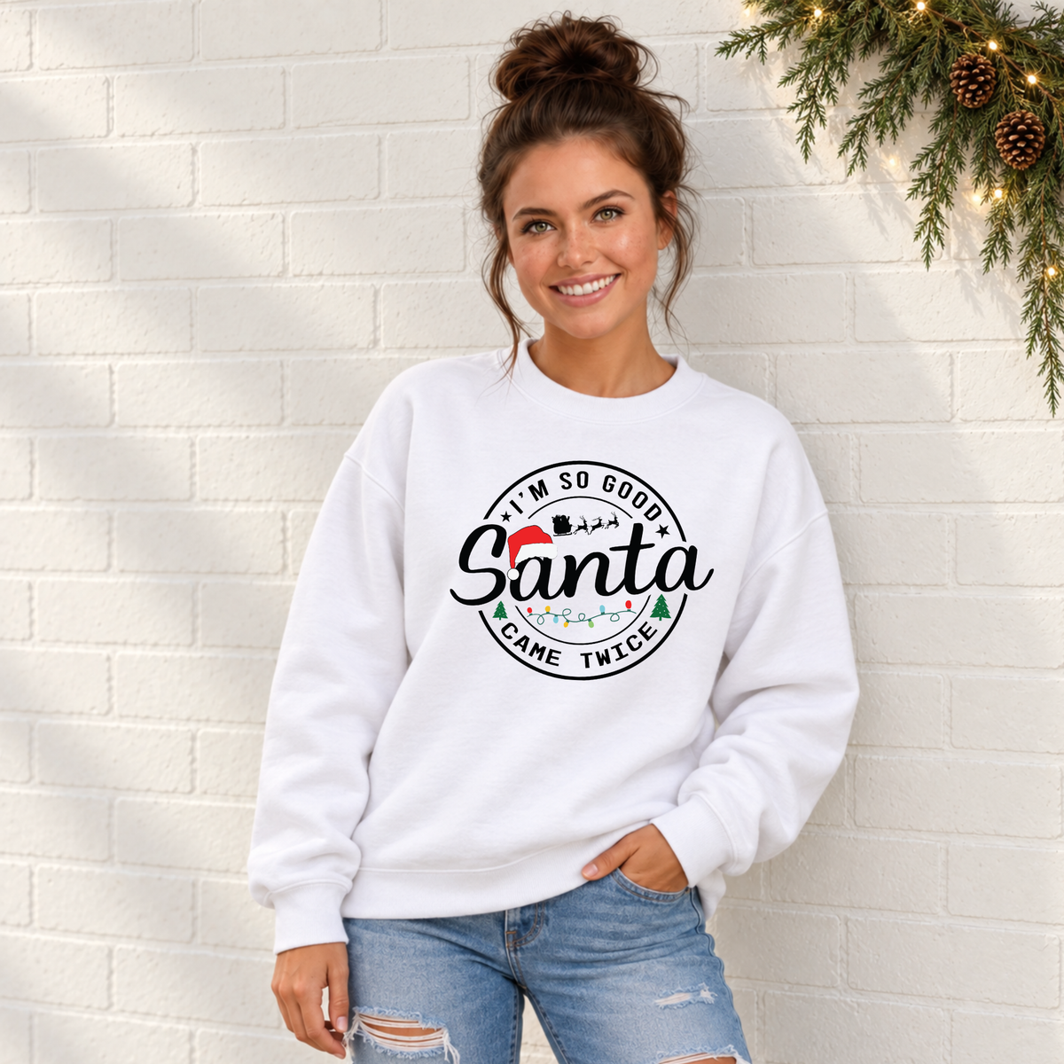 Woman wearing a white sweatshirt with I'm So Good Santa Came Twice Christmas design against a white brick wall