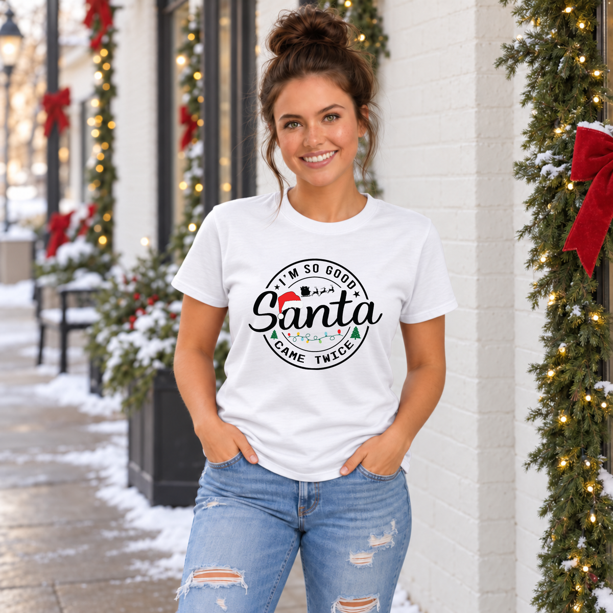 Woman wearing a white T-shirt with I'm So Good Santa Came Twice Christmas design outside a snowy holiday storefront