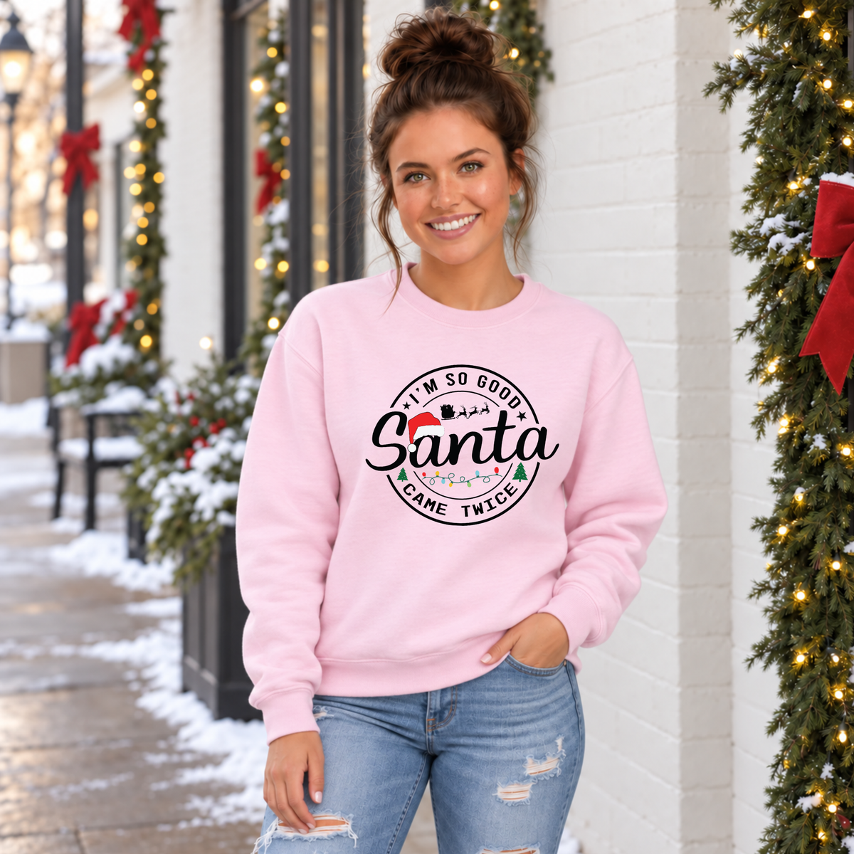 Woman wearing a light pink sweatshirt with I'm So Good Santa Came Twice Christmas design outside a snowy holiday storefront