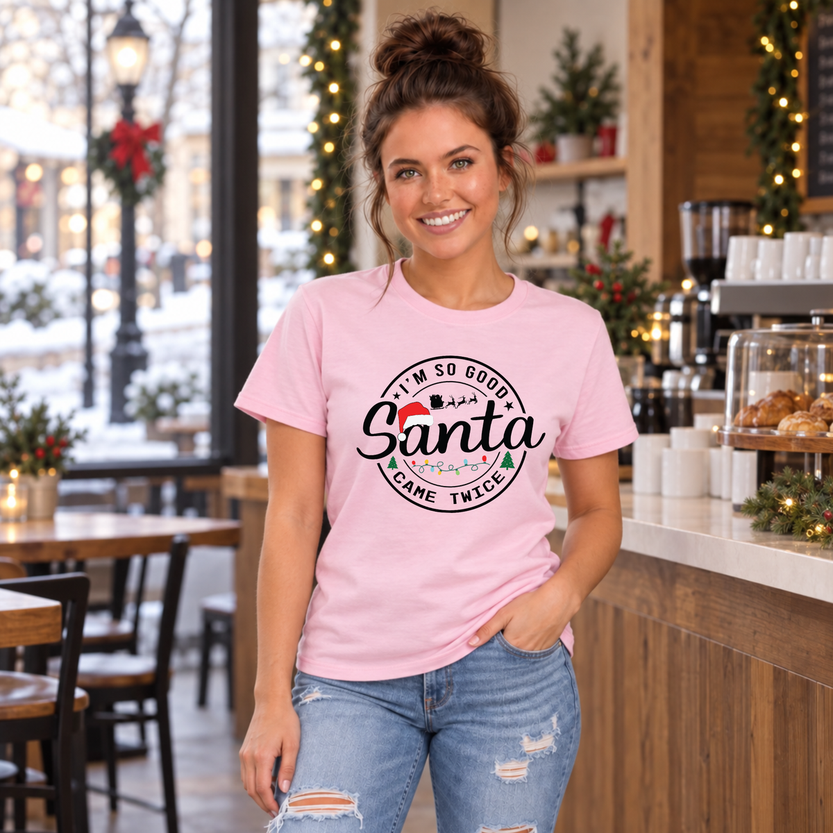 Woman wearing a light pink T-shirt with I'm So Good Santa Came Twice Christmas design in a festive café