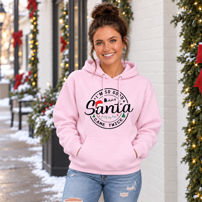 Woman wearing a light pink hoodie with I'm So Good Santa Came Twice Christmas design outside a snowy decorated storefront