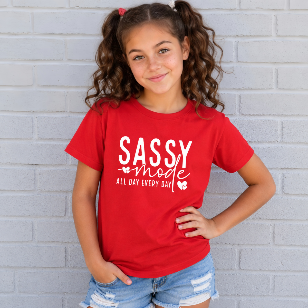 Youth girl wearing a red Sassy Mode T-shirt with white lettering against a light brick wall.