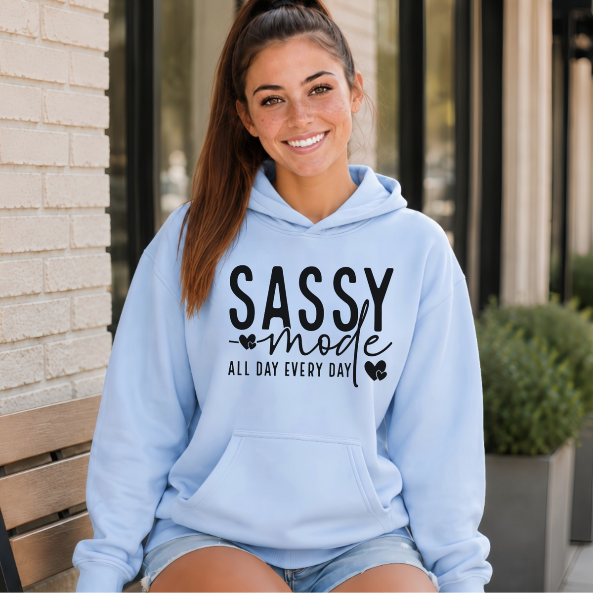 Woman wearing a light blue Sassy Mode hoodie with bold black lettering outside a storefront.