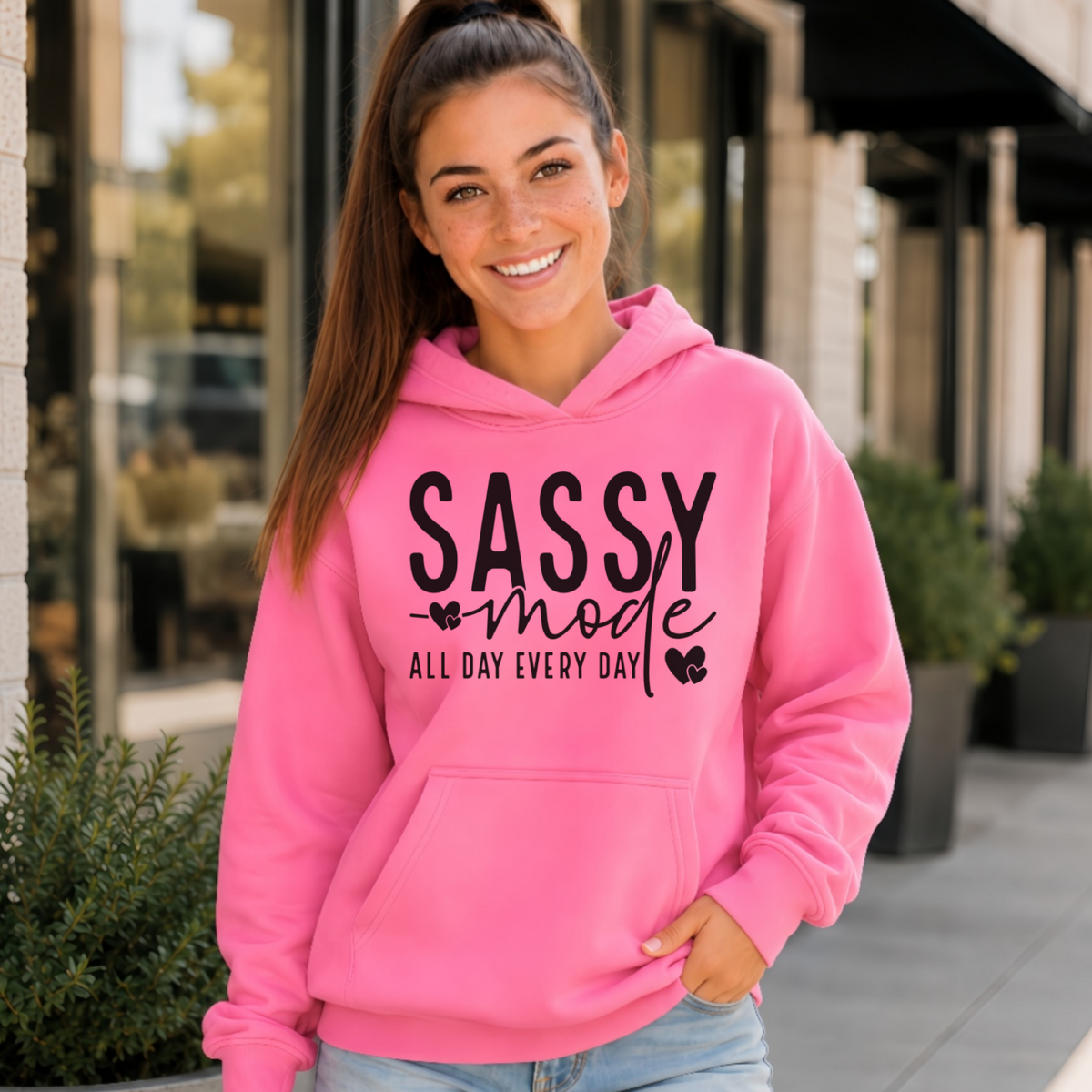 Woman wearing a bright pink Sassy Mode hoodie with bold black lettering outside a storefront.