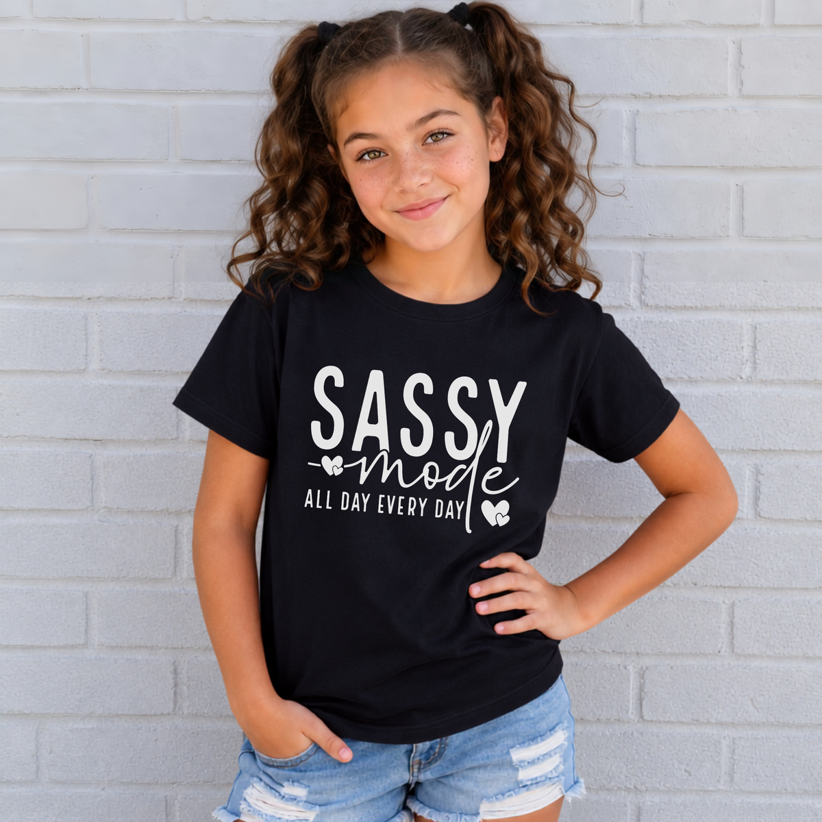 Youth girl wearing a black Sassy Mode T-shirt with white lettering against a light brick wall.