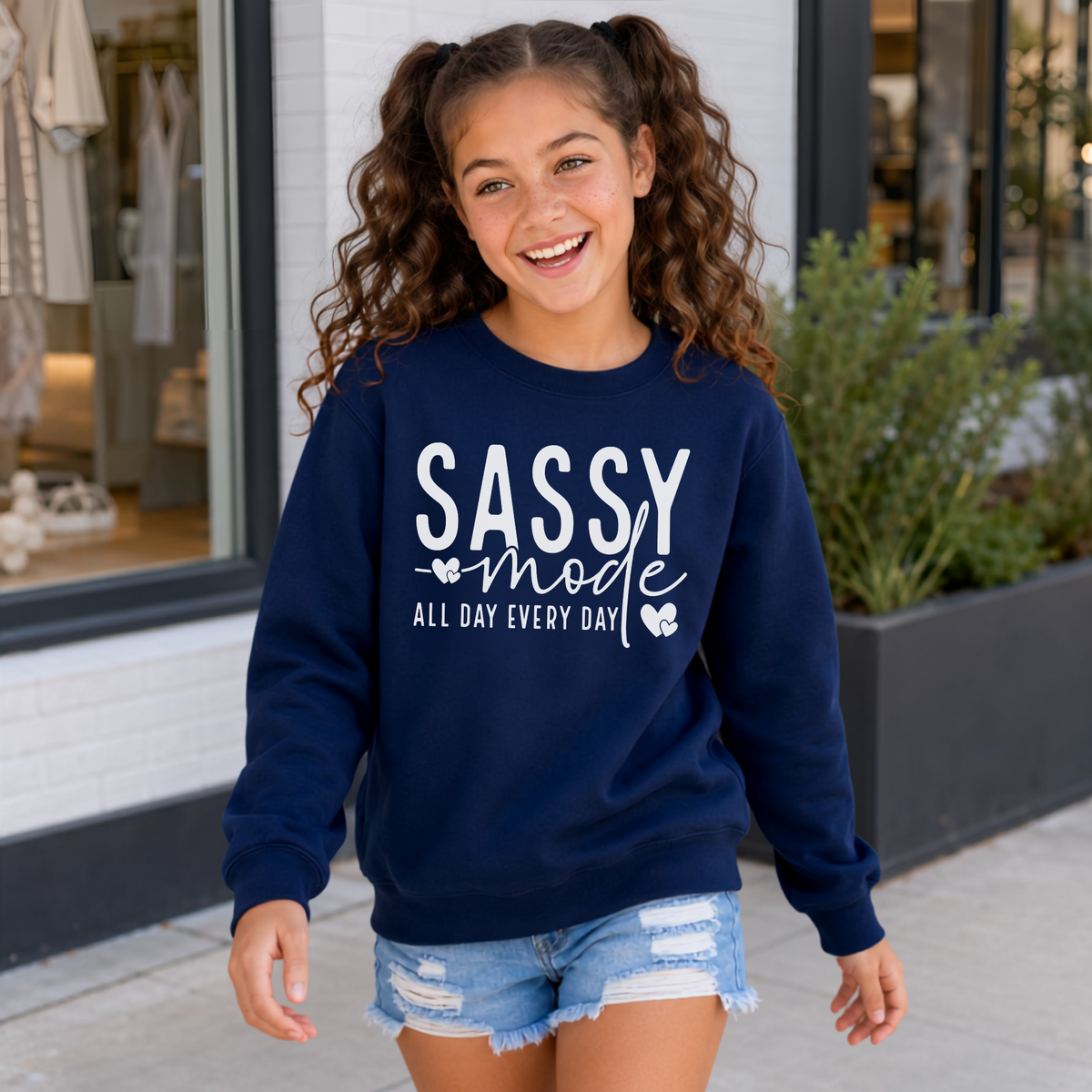 Youth girl wearing a navy Sassy Mode sweatshirt with white lettering outside a storefront.